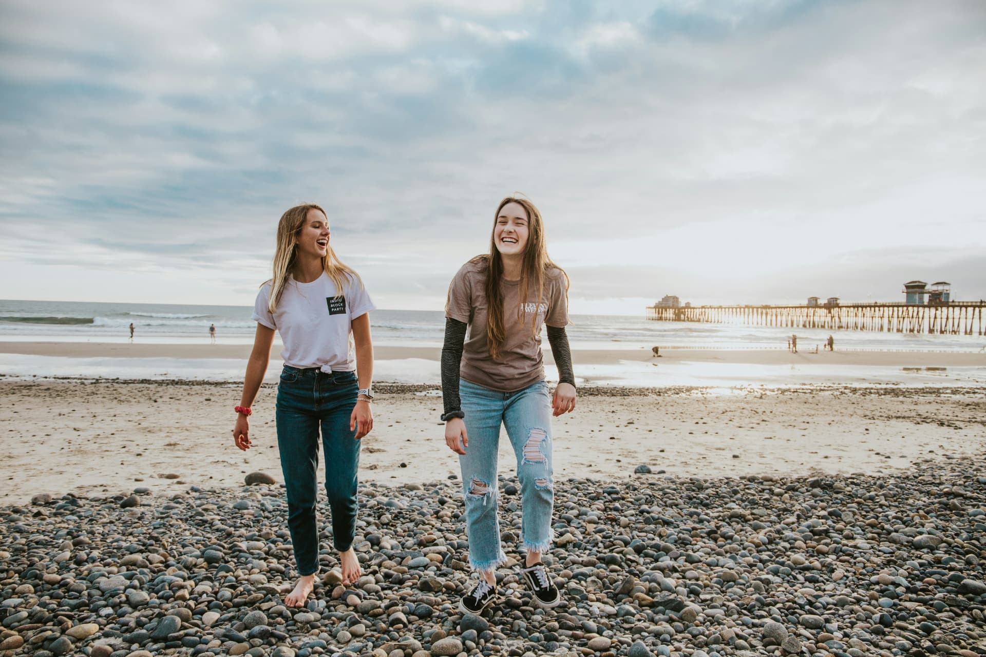 Two young women laughing together on a rocky beach at sunset.